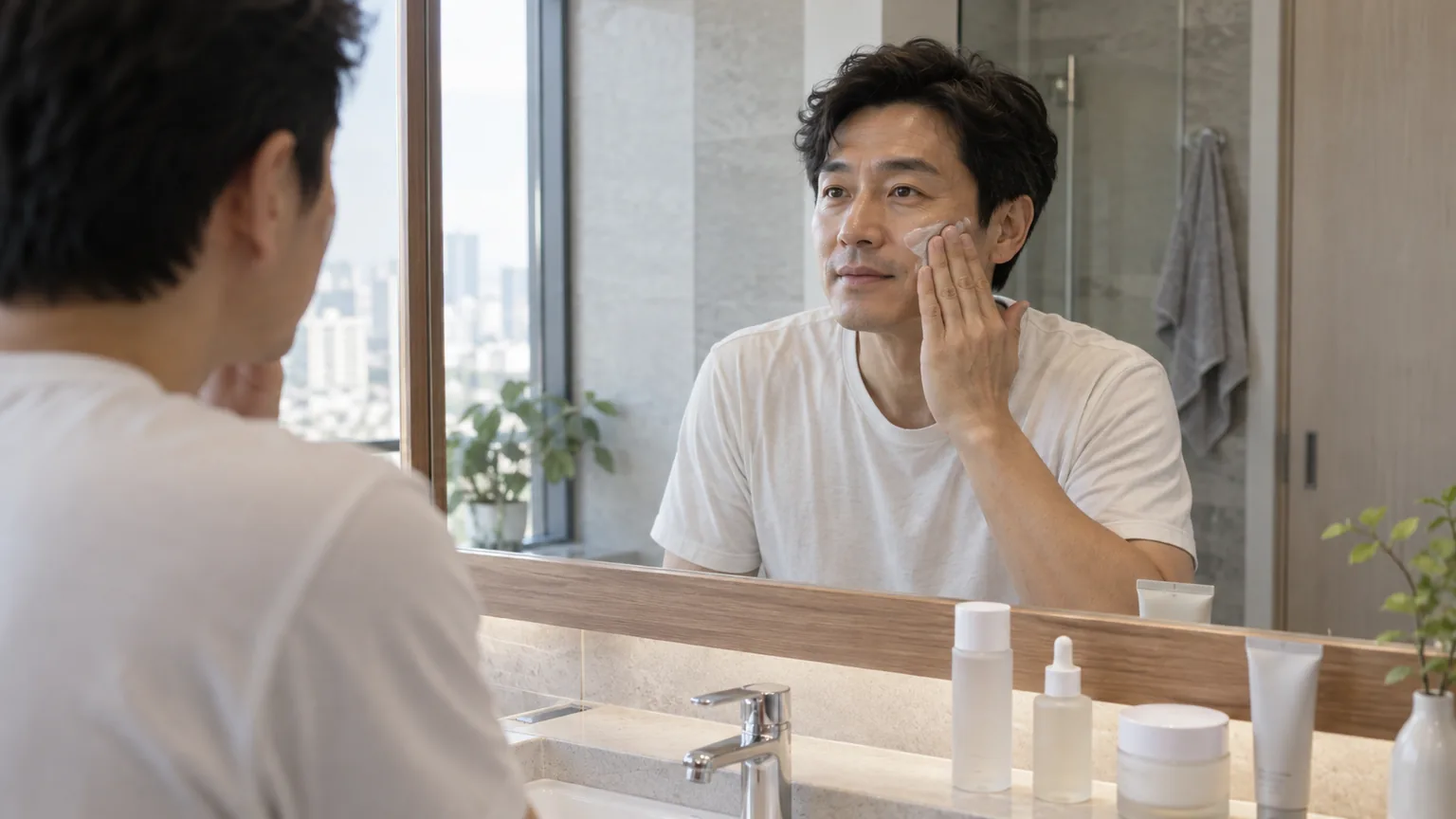 A Korean man in his 40s applying moisturizer at a bathroom mirror beside four unbranded skincare containers